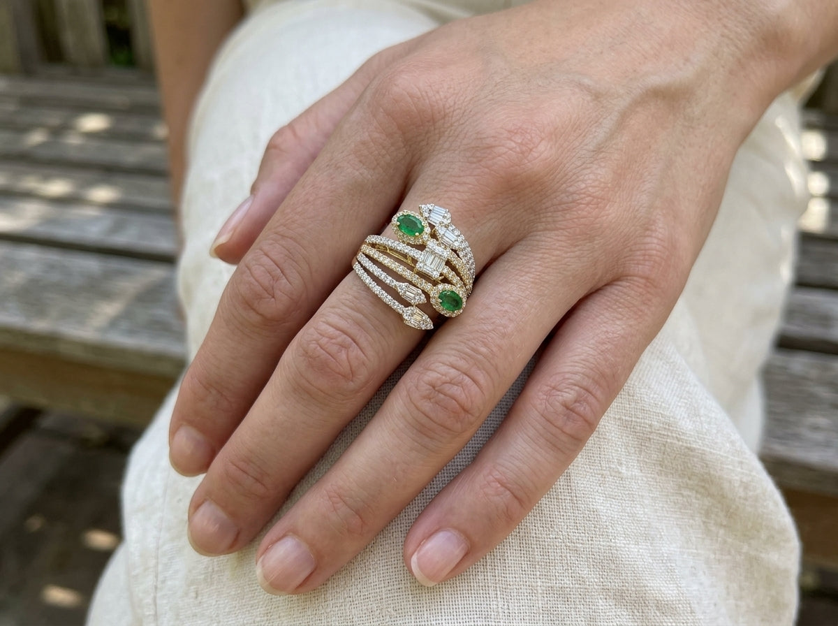 A close-up of a person's hand wearing a stack of three gold rings with green emerald-cut stones and clear pavé accents, resting on a cream linen surface.